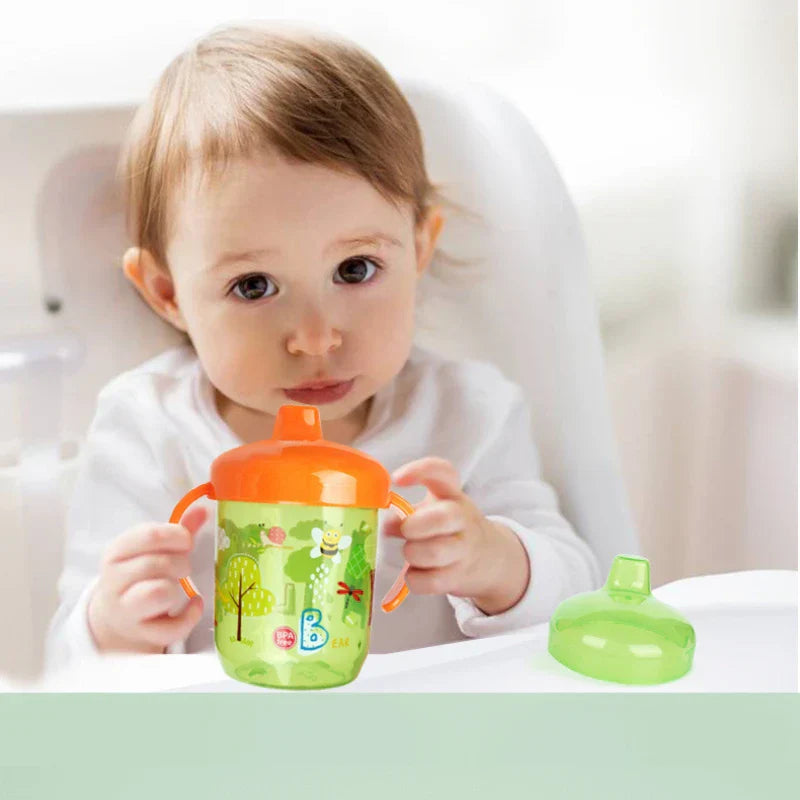 Bébé assis dans une chaise haute buvant avec un gobelet d'apprentissage coloré à anses, couvercle vert à côté.