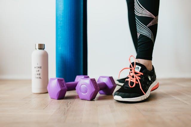 Femme en tenue de sport avec haltères violets, tapis de yoga et gourde sur parquet