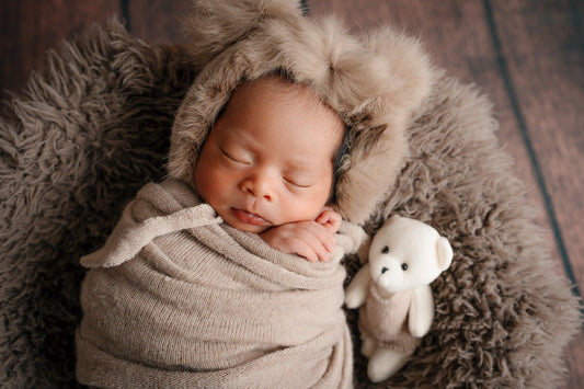 Nouveau-né endormi sur plaid marron, bonnet en fourrure, doudou peluche à côté
