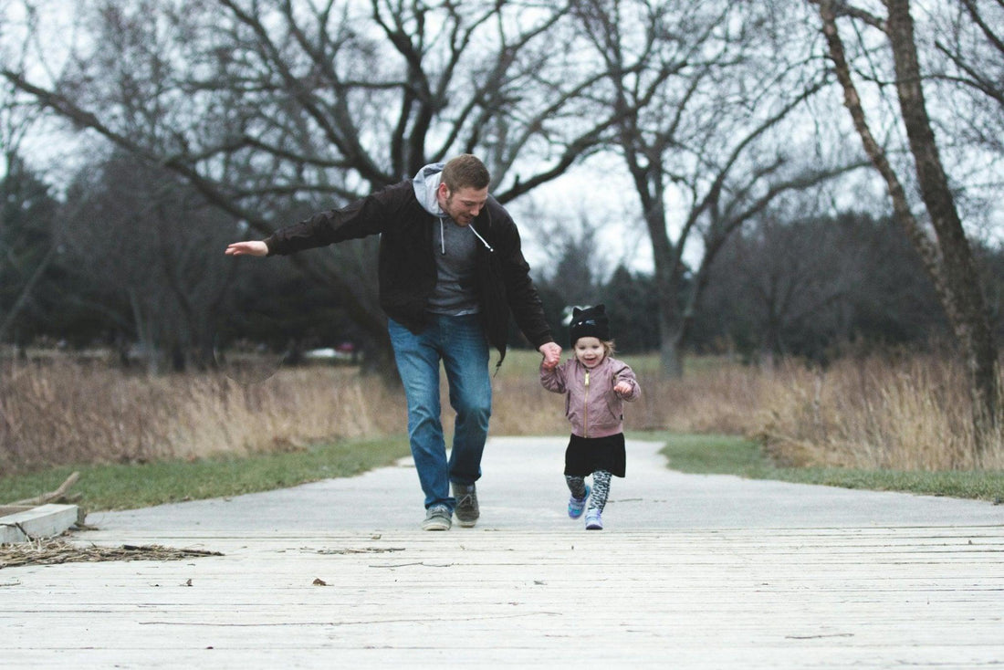 Papa promenant sa fille souriante en automne sur un chemin bordé d’arbres sans feuilles.