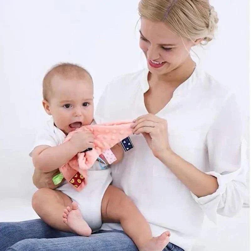 Bébé assis sur les genoux d'une femme, tenant un doudou sensoriel rose à étiquettes colorées.