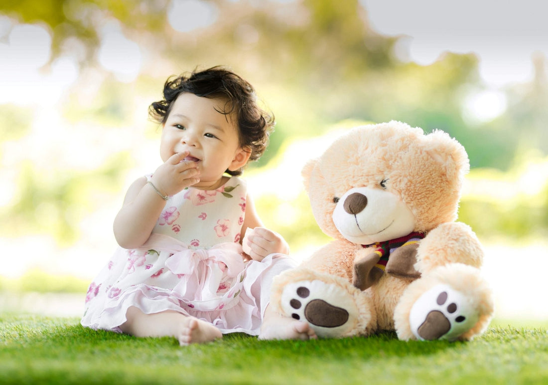 Bébé assis sur l’herbe avec une robe à fleurs, à côté d’un doudou peluche ourson.