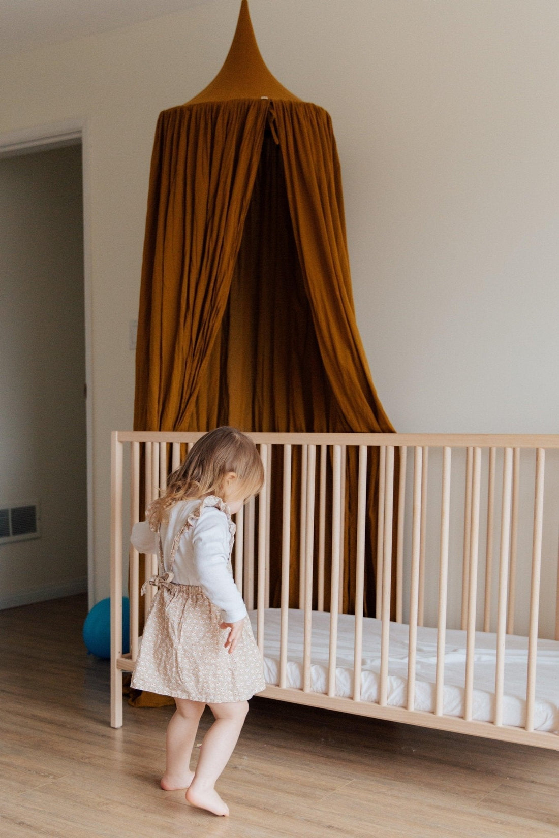 Petite fille près d’un lit bébé en bois avec ciel de lit moutarde dans une chambre d’enfant.