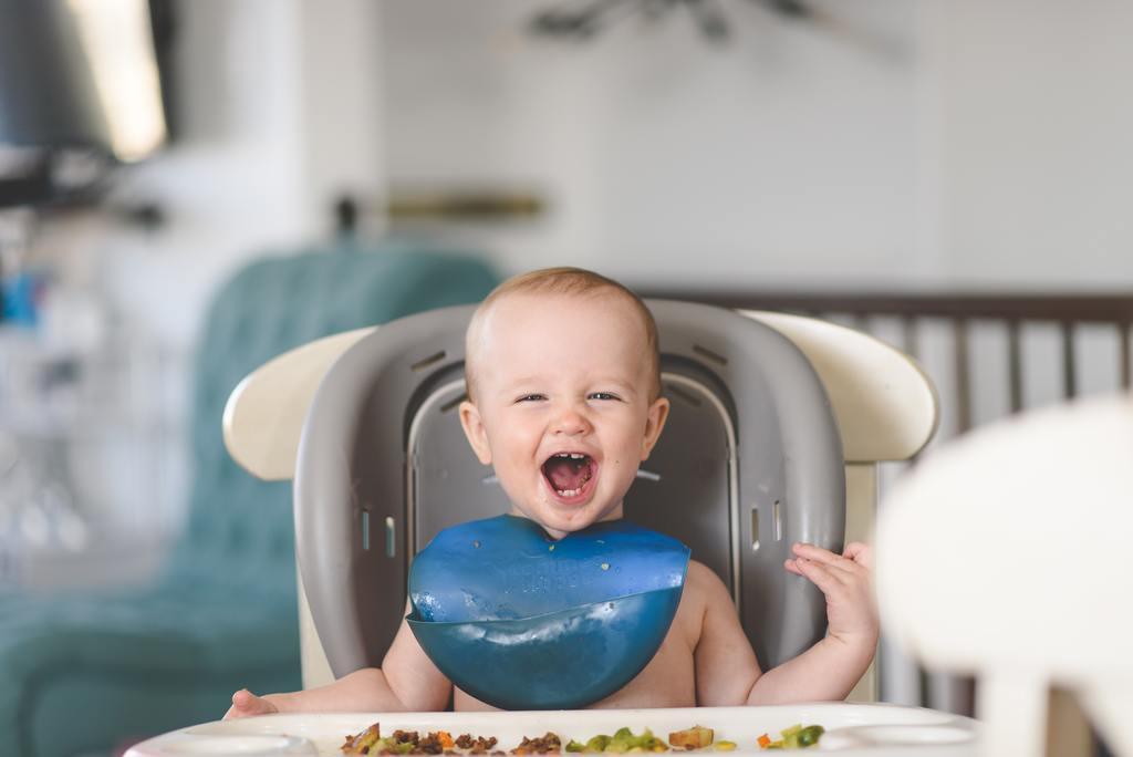Bébé souriant dans une chaise haute, bavette bleue, repas de diversification sur la table.